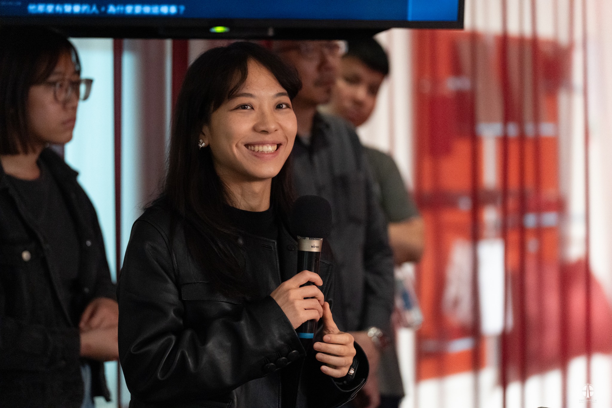 A photograph of artist Lee Tzu Tung speaking into a microphone at an indoor event. They are smiling brightly, with dark shoulder-length hair, wearing a black leather jacket, delicate earrings, and a watch. Other attendees stand in the background, along with a blue screen overhead and red curtains.