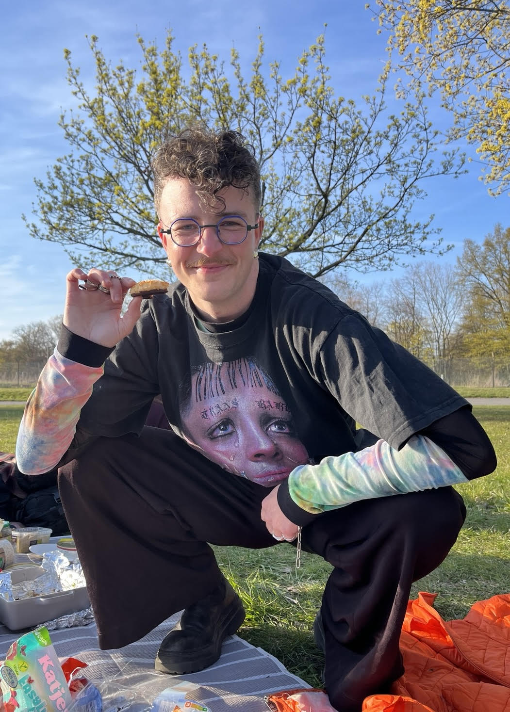 Ren is squatting in the park, a tree emerges from behind them, it’s a picnic scene. They are holding a cookie and wearing a t-shirt that reads ’TRANS BABE’ across the top of it. Ren is a white, trans*masculine person wearing silver heart earrings, colorful glasses and lots of rings.
