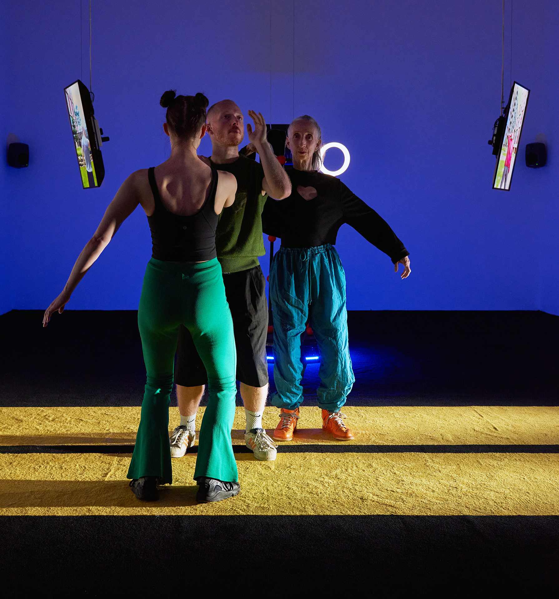 three characters standing on a stage. Nina Davies - Precursing performance. Photo by Jonathan Bassett, 2023.