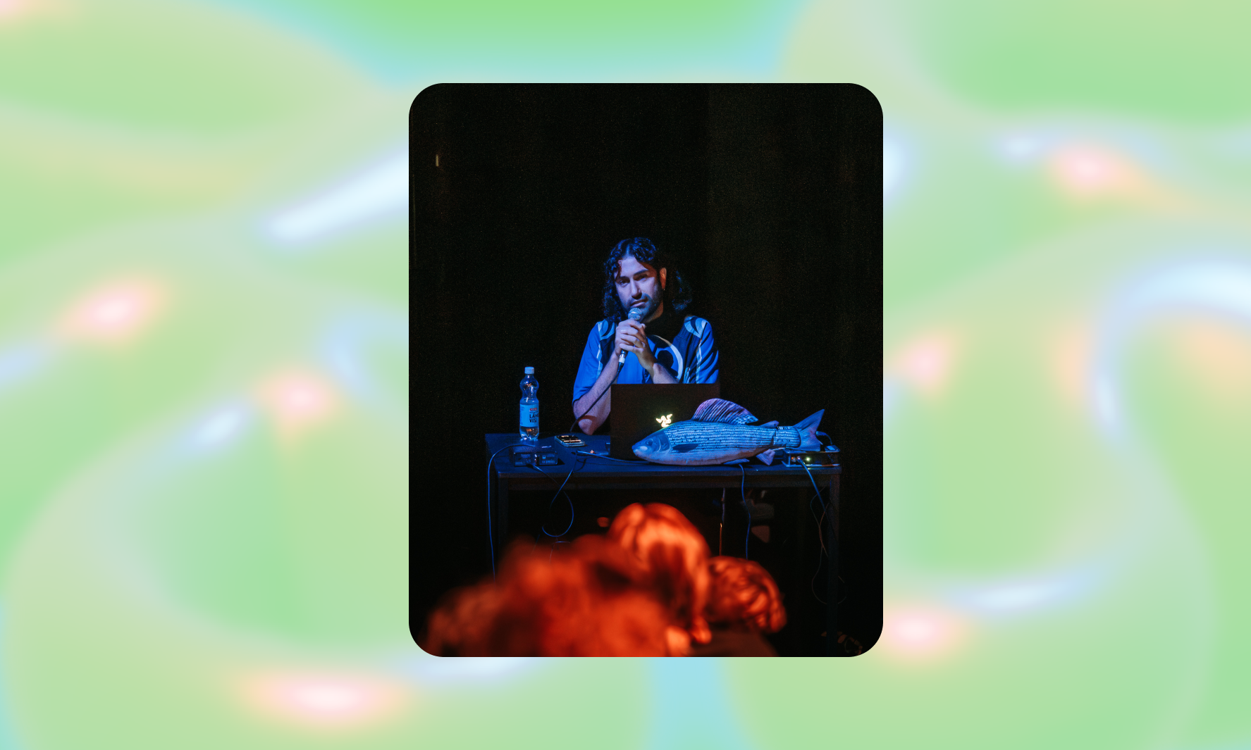 Harold Hejazi holding a microphone, sitting behing a laptop. There is a fish character on the table. Room is dark, there is blue light on Harald.