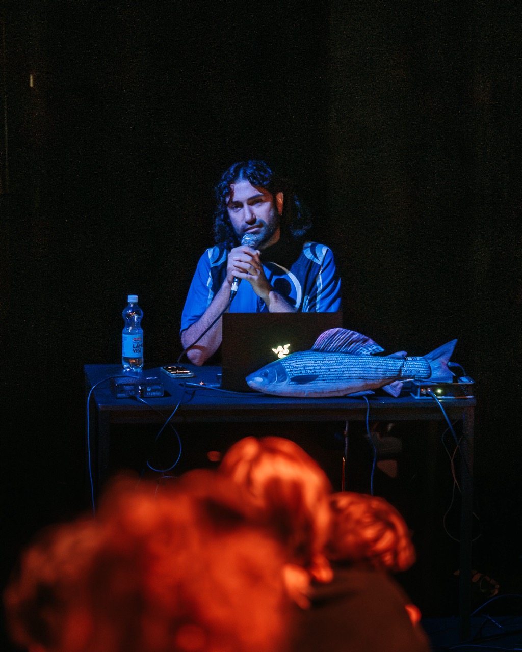 Harold Hejazi holding a microphone, sitting behing a laptop. There is a fish character on the table. Room is dark, there is blue light on Harald.